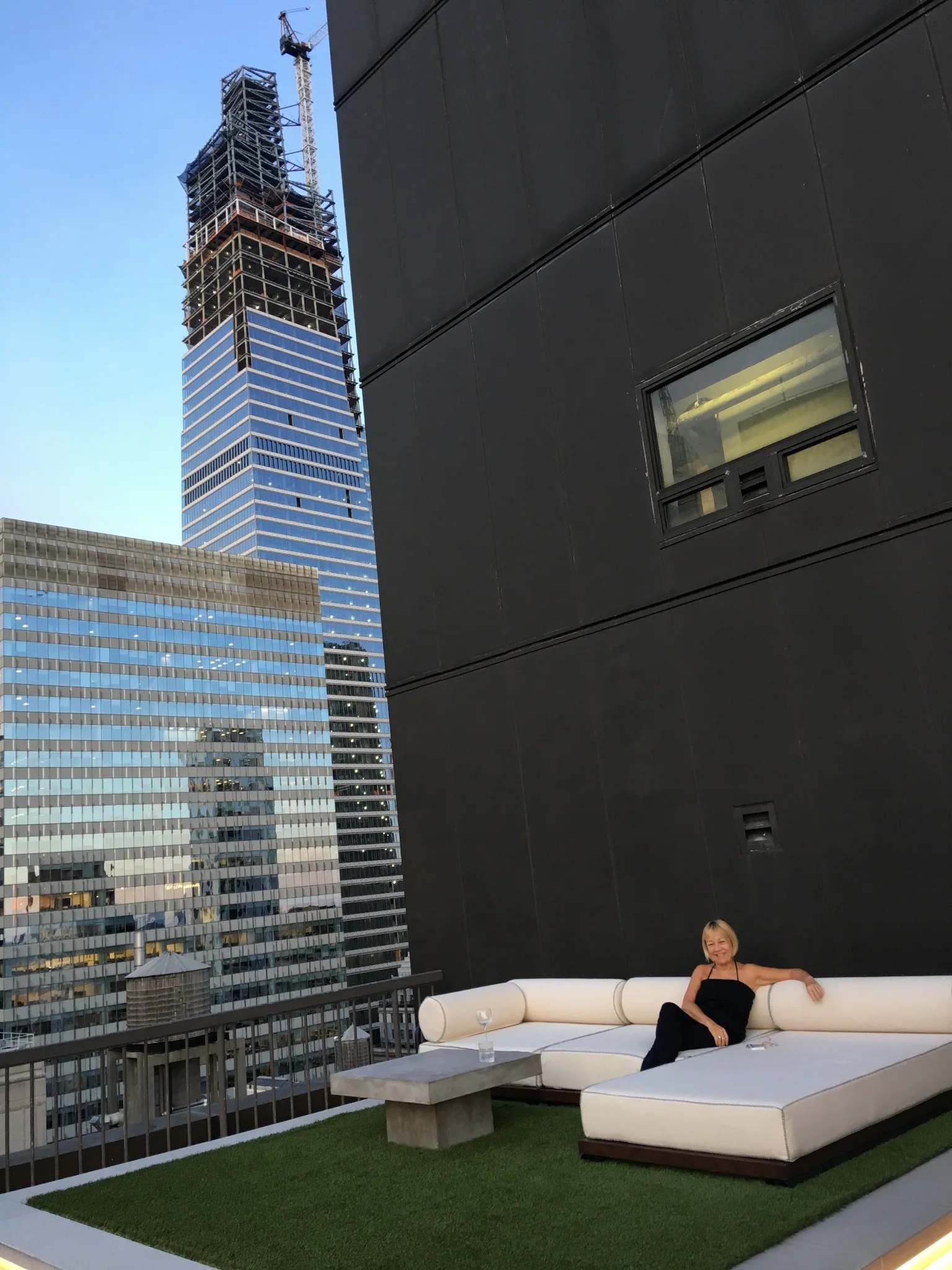 Cindy Gallop seated on a white outdoor sofa on the Sky Apartment terrace, with Manhattan buildings behind her.