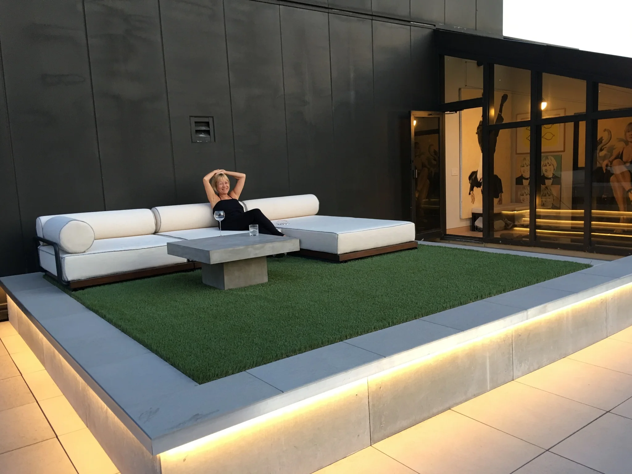 Cindy Gallop relaxing on the Sky Apartment terrace beside a white sofa, greenery, and illuminated steps.